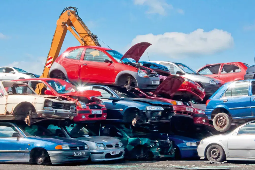 A crowded scrapyard filled with stacked, damaged and discarded cars, with an orange excavator lifting vehicles in the background under a bright blue sky.