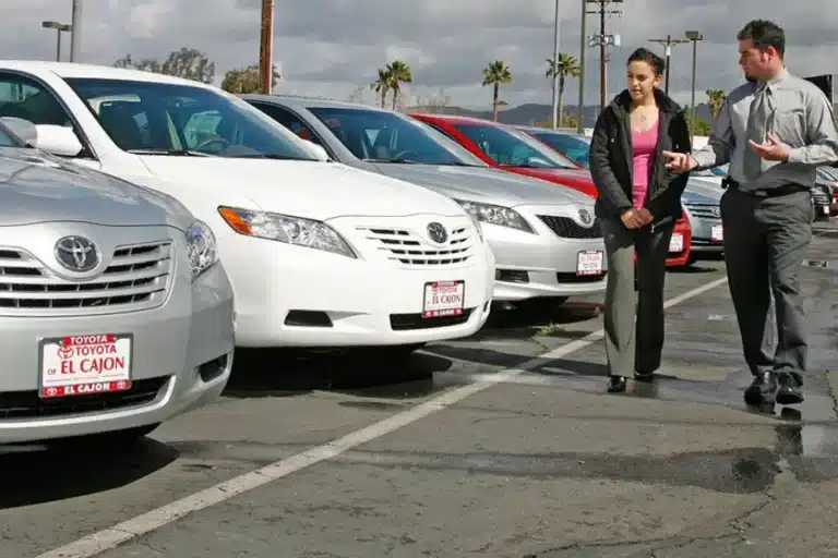 A man and woman discuss cars at a dealership, standing beside a row of mostly white Toyota vehicles in an outdoor parking lot.
