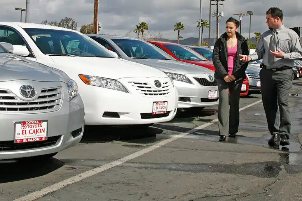 A man and woman discuss cars at a dealership, standing beside a row of mostly white Toyota vehicles in an outdoor parking lot.